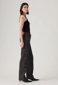 Black denim jeans with decorative studded accents down the side. High-waisted fit, wide leg design, paired with a fitted black tank top and heeled boots.