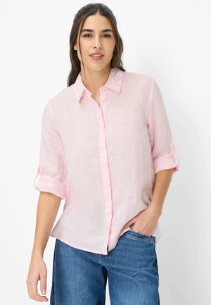 Mujer de cabello largo y oscuro, con una camisa rosa claro de botones y mangas remangadas, y jeans azules, de pie con una mano en el bolsillo.