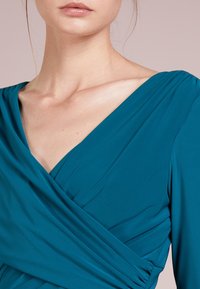 Teal omslagsklänning gjord av mjuk, elastisk tyg med djup V-ringning och samlade detaljer över bysten.