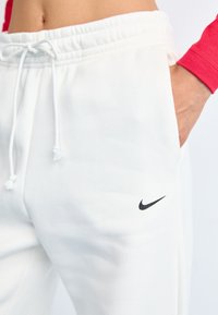 Bílé tepláky vyrobené z měkkého materiálu, s elastickým pasem a šňůrkou, a s malým černým logem Nike na boku.