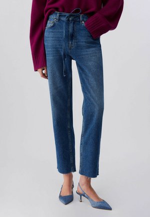 Ženska, oblečena v pridelane modre kavbojke z ravnimi hlačnicami in zunanji robovi, s pasom, ki je zavezano, bordo pulover in modre petke z odprto peto z okraski.