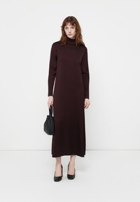 Robe longue bordeaux avec col roulé, manches longues et silhouette droite. Portée avec des talons hauts noirs et un sac à main noir.