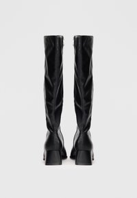 Une paire de bottes noires en cuir, hautes avec des talons blocs, vue de l'arrière sur un fond blanc uni.