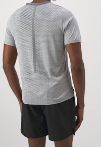 Šedé sportovní tričko s texturovaným vzorem, krátkými rukávy a reflexním vertikálním akcentem na zádech, ve spojení s černými šortkami.