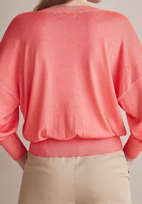 Kobieta w luźnym koralowym swetrze z szerokimi rękawami, włożonym w jasnobeżowe spodnie o wysokim stanie, pokazana z tyłu.