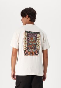Camiseta de algodón crema con un colorido estampado gráfico en la parte posterior, que incluye setas, un caracol y elementos de texto. Manga corta, corte relajado.