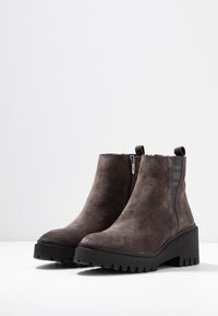 Šedé semišové kotníkové boty s robustní černou podrážkou, bočním zipem a elastickými panely pro snadné obouvání. Texturovaný svršek a pevná konstrukce.