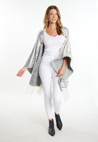Poncho aperto in maglia grigia e crema con motivo geometrico, bordo frangiato, indossato sopra una maglietta bianca e pantaloni bianchi, abbinato a stivaletti neri.