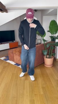 Juodas zip-up purvasuknis su baltais dryželiais, derinamas su mėlynomis plačiomis kelnėmis ir baltais sportbačiais, įrengtoje šiuolaikinėje patalpoje.