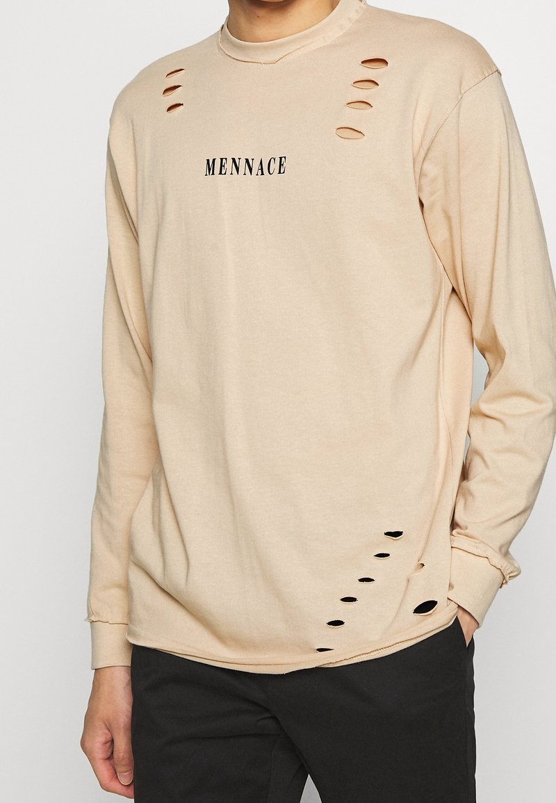 Beige košile s dlouhým rukávem, s výřezy na ramenou a na spodním lemu, s černým textem, který říká "MENNACE." Měkká textilie.