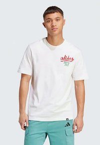 Jauns vīrietis, tērpts baltā Adidas T-kreklā ar sarkano un zaļo logo, kā arī tumšzila krāsas cargo biksēs ar melno Adidas logo, stāv uz gaiša fona.