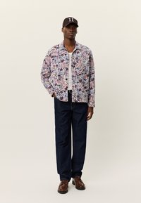 Ελαφρύ πουκάμισο-ζακέτα με λουλουδάτο σχέδιο, πολύχρωμο, με γιακά και κλείσιμο με κουμπιά. Σκούρο μπλε τζιν παντελόνια και καφέ παπούτσια.