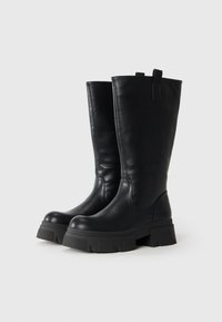 De grandes bottes noires en cuir avec des semelles épaisses à plateforme et des tirettes, présentées côte à côte sur un fond clair.