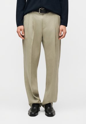 Hombre vestido con pantalones plisados beige, cinturón negro y zapatos de vestir de cuero negro, de pie contra un fondo liso.