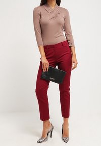 Frau trägt ein mauvefarbener Wickel-Oberteil, burgunderrote knöchellange Hose, Schlangenleder-Muster hohe Absätze und hält eine schwarze Clutch.