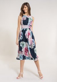 Frau in einem ärmellosen, marineblauen Midikleid mit großem, pinkfarbenem, blauen und weißen Blumenmuster, die mit einem Bein nach vorne steht und lächelt, dabei nudy-farbene Absätze trägt.