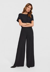 Czarna satynowa bluzka z krótkimi rękawami, w połączeniu z szerokimi czarnymi spodniami. Spodnie mają gładką teksturę i subtelny detal w postaci rozcięcia z przodu.