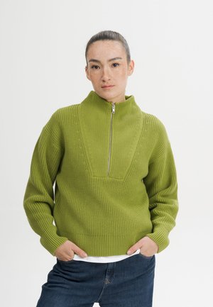 Vrouw met haar naar achteren gebonden, een groene gebreide trui met halve rits en blauwe spijkerbroek dragend, staand met handen in de zakken tegen een witte achtergrond.