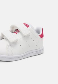 Vit velcro-sneaker med perforerad design, rosa detaljer på hälen och logotypen, samt en gummisula. Har ett slätt syntetmaterial.