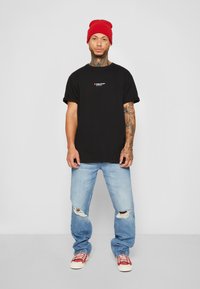 T-shirt preta de algodão com um pequeno logótipo gráfico, combinado com jeans claros desfiados e ténis vermelhos. Gorro de lã vermelho na cabeça.