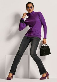Lila gerippter Rollkragenpullover, schwarze enge Jeans mit fransigem Saum, lila Wildleder-Heels und eine schwarze strukturierte Handtasche. Runde Sonnenbrillen runden das Outfit ab.