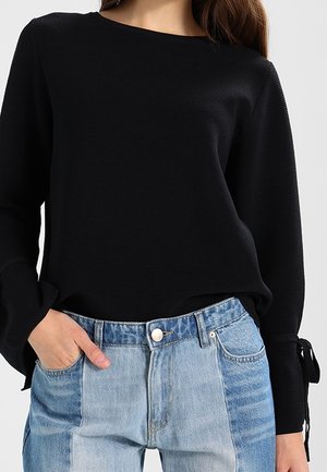 Top lungo a maniche ribbed nere con scollatura rotonda, dotato di polsini con fiocco. Abbinato a jeans di denim azzurro chiaro a patchwork con chiusura a bottone.