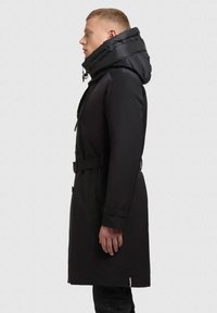 Juodas paminkštintas ilgas paltas su aukštu apykakle, turintis diržo juosmeniui, mygtukus užsegimui ir tekstūruotus akcentus šilumai ir stiliui.