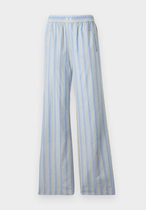 Femme debout les mains dans les poches, portant un blazer en cuir noir, un top blanc, un pantalon ample bleu rayé et des chaussures noires sur fond blanc.