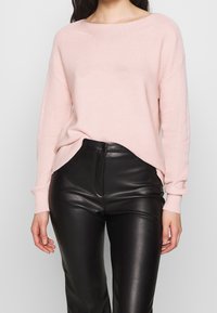 Femme portant un pull en maille rose clair rentré dans un pantalon en cuir noir taille haute, debout devant un fond clair uni.