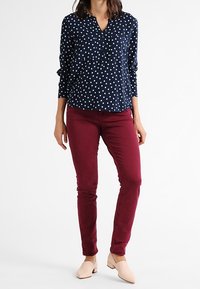 Chemise à pois marine avec une coupe décontractée, décolleté en V ; associée à un pantalon slim bordeaux ; des ballerines beiges complètent la tenue.