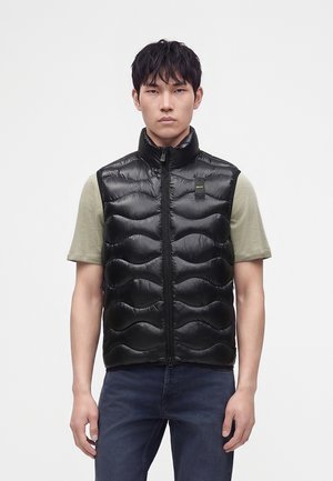 Jeune homme portant un gilet matelassé noir sans manches sur une chemise manches courtes vert clair et un jean bleu, debout devant un fond uni.