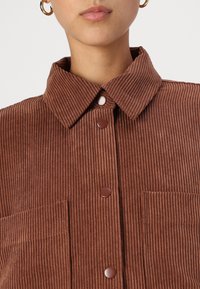 Camicia in corduroy marrone con colletto, tre bottoni marroni e due tasche a toppa. Il tessuto ha una texture a coste.