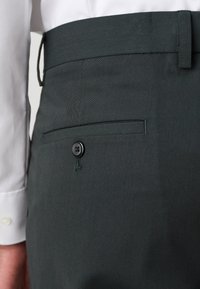 Dark green pants with a smooth texture, featuring a side pocket with a black button closure. White collared shirt partially visible.