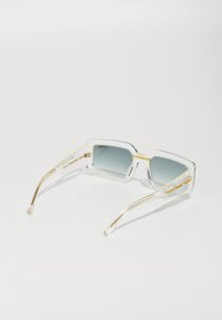Ochelari de soare cu ramă transparentă și lentile gri cu gradient, temple accentuate cu aur și o formă geometrică. Brațele texturate prezintă un model marmorat.