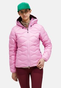 Giacca imbottita rosa chiaro con design a ondulato, zip frontale e cappuccio regolabile. Indossata sopra una maglia beige e pantaloni bordeaux.