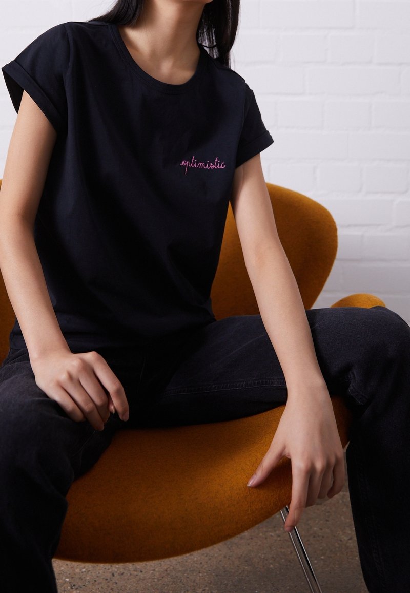 Svart bomull t-shirt med rullade ärmar och rosa broderad text som säger "optimistisk." Bärs med mörka jeans, sittande på en orange stol.
