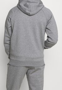 Sudadera gris con capucha ajustable, puños y dobladillo acanalados, con una textura suave y mangas raglán. Se lleva con pantalones de chándal grises a juego.