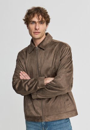 Uomo con capelli ricci che indossa una giacca marrone con cerniera e jeans blu, in piedi con le braccia incrociate davanti a uno sfondo semplice.
