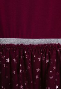 Bordeauxrotes Kleid mit einem taillierten Oberteil und einem mehrlagigen Rock. Der Rock ist mit silbernen Sternmustern versehen und hat einen glitzernden silbernen Taillenbund.