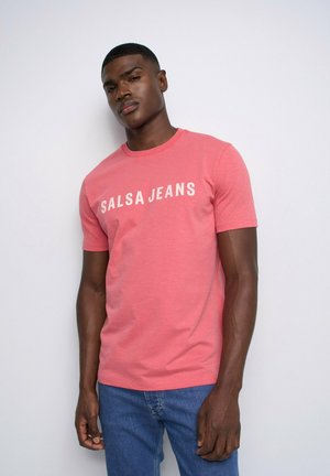 Hombre con una camiseta rosa "Salsa Jeans" y vaqueros azules de pie frente a un fondo blanco liso.