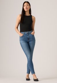 Schwarzes geripptes Tanktop, hochtaillierte hellblaue Skinny-Jeans mit fünf Taschen und schwarze Ballerinas. Die Jeans zeigen einen leichten Farbverlauf zum Saum hin.