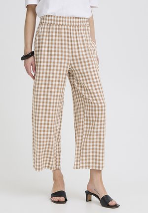 Personne portant un pantalon large à carreaux beige et blanc avec une taille élastique, associé à des sandales noires à talons et un haut blanc.