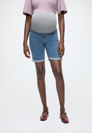 Persoană însărcinată purtând un top roz, o bandă maternă gri pentru burtă, pantaloni scurți din denim suflecați și pantofi roșii Mary Jane, stând pe un fundal alb.