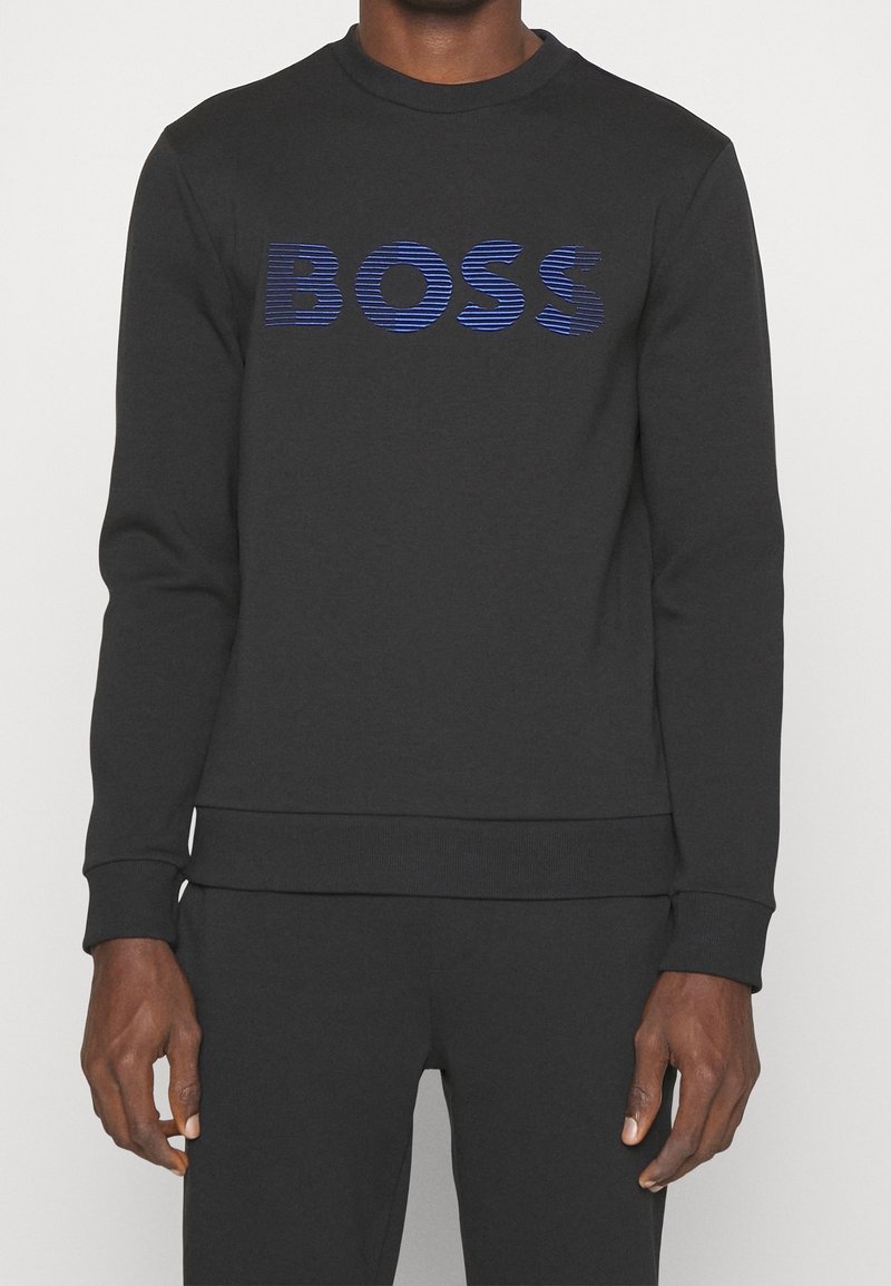 Sweat-shirt noir à manches longues avec inscription « BOSS » rayée en bleu, porté par une personne debout avec les mains détendues le long du corps.