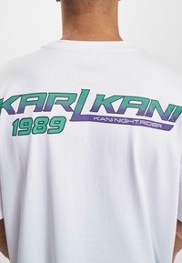 Balta medvilninė marškinė su dideliu grafikiniu spaudiniu ant nugaros, kuriame užrašyta "KARL KANI 1989" žaliai ir purpuriškai, o žemiau parašyta "KANI NIGHT RIDER".
