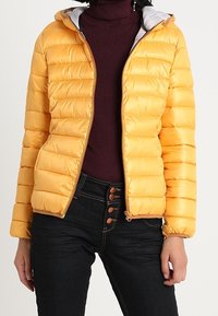 Femme portant une veste matelassée jaune vif par-dessus un pull à col roulé bordeaux et un jean foncé à bouton, se tenant les mains détendues sur les côtés.