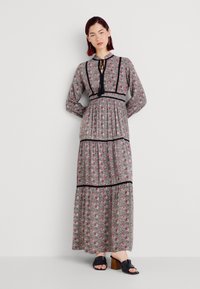 Robe maxi à motifs floraux en gris avec des accents noirs, dotée de manches longues, d'un col à nouer et d'un design à volants. Portée avec des sandales à talons noires.