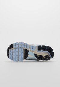 Suela de una zapatilla con patrones de tracción en goma de color azul claro, negro y beige sobre un fondo blanco liso.