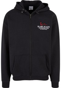 Sudadera con cremallera negra que presenta un bolsillo frontal, puños y dobladillo acanalados, y un logo bordado en blanco y rojo. Tejido suave y texturizado.