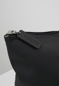 Pochette noire en nylon avec une surface texturée, dotée d'une fermeture zippée sur le dessus et d'une étiquette en caoutchouc portant le logo Lacoste attachée à la fermeture éclair.